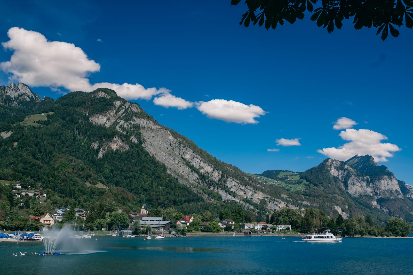Blick über den Walensee auf Weesen: das Dorf mit der Kirche am Ufer, dahinter die bewaldeten Felswände, davor ein Kursschiff und die Springbrunnenfontäne
